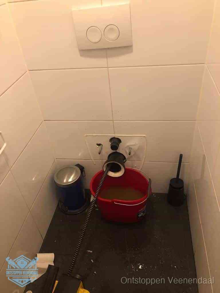 Een ontstopte wand-wc met verwijderde toiletpot waar een ontstoppingsveer in de wanduitgang verdwijnt, het achterhalen waar de verstopping zit in Veenendaal