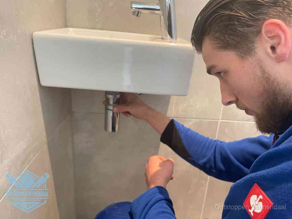Een monteur draait met de hand de zwanenhalssifon onder een fonteinwasbak in de toiletruimte los om de bron van rioollucht in Veenendaal te vinden