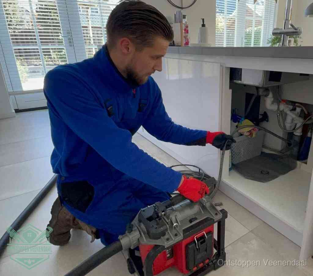 Een monteur met rode handschoenen knielt in een keuken en voert een elektrische ontstoppingsveer in tijdens het professioneel afvoer ontstoppen in Veenendaal