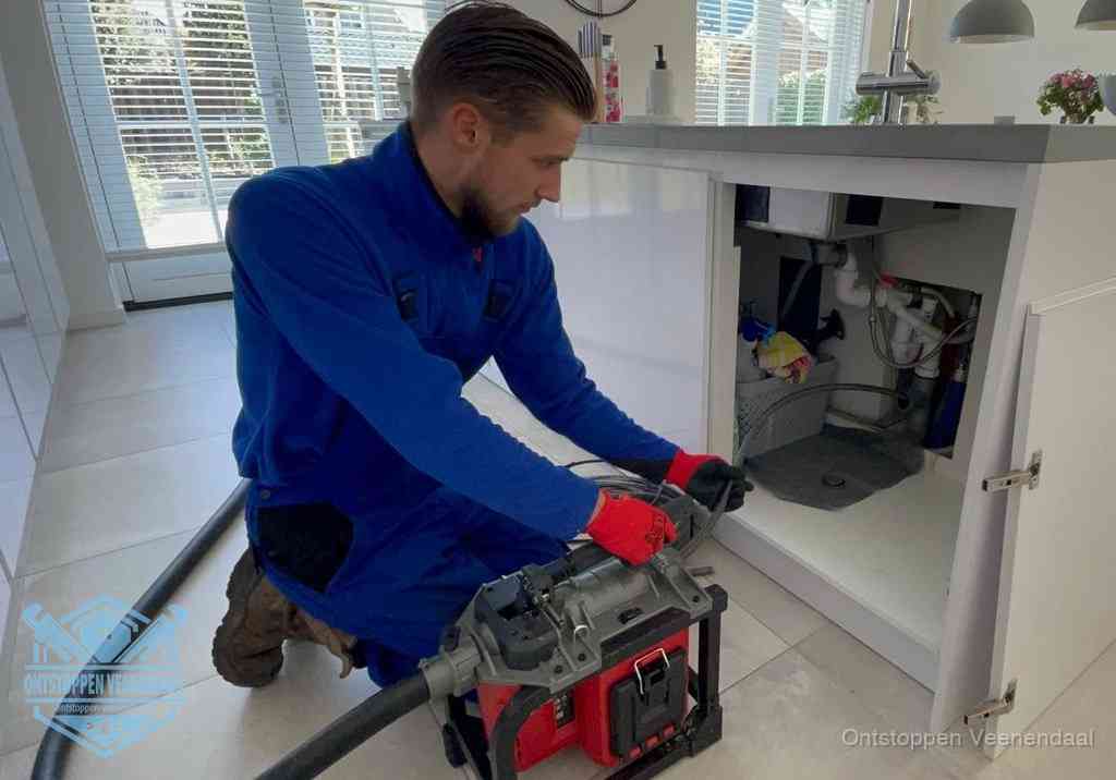 Een monteur voert een professionele elektrische ontstoppingsmachine in de afvoer onder de spoelbak tijdens het gootsteen ontstoppen in Veenendaal